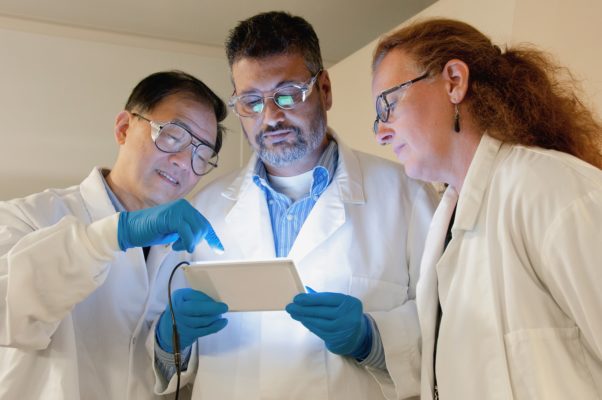 Three scientists in white lab coats looking at a tablet.